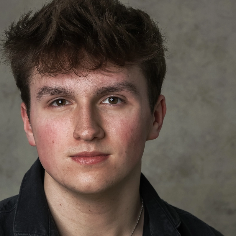 A young man, Andy Norbuta, with short, tousled brown hair and fair skin faces the camera. He has brown eyes, a neutral expression, and is wearing a dark jacket over a shirt. The background is a textured gray.