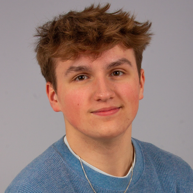 A young person with short, tousled light brown hair and fair skin smiles softly at the camera. They wear a light blue textured sweater and a silver necklace. The plain, light gray background gives this Andy Norbuta portrait a simple and warm feel.