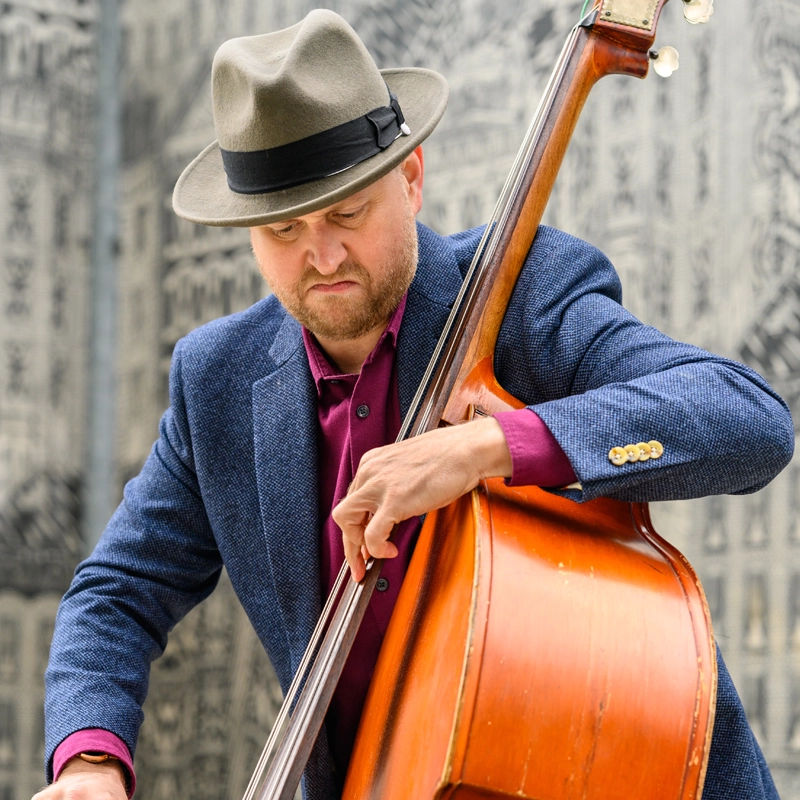Michael O'Brien, in a gray fedora, blue blazer, and magenta shirt, plays a double bass. He looks down intently at the strings while abstract patterns in shades of gray and black blur the background.