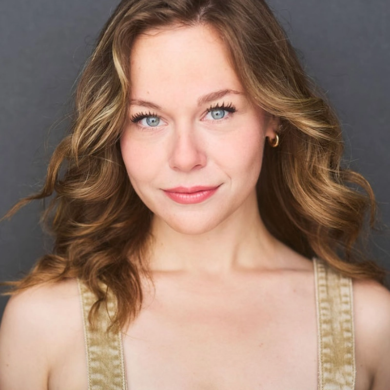A woman, McKenna OGrodnik, with wavy light brown hair and blue eyes looks directly at the camera, smiling slightly. She wears a sleeveless beige top and gold hoop earrings, set against a plain dark gray background for a professional portrait feel.