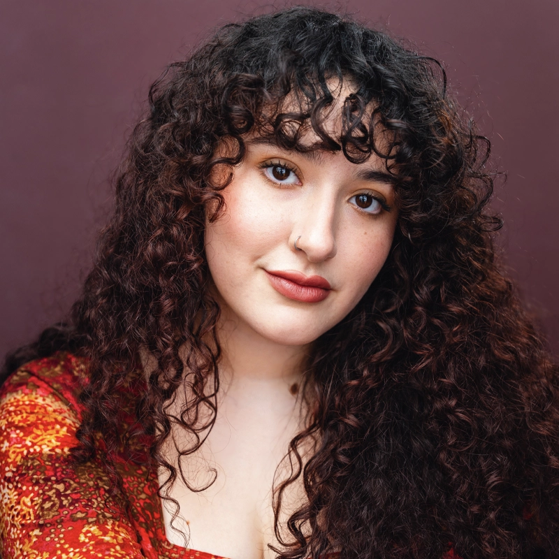 Corinne O'Reilly, with long, dark, curly hair and bangs, poses against a purple background. She has a light complexion, brown eyes, natural makeup with rosy lipstick, wears a red patterned top, and looks at the camera with a soft smile.