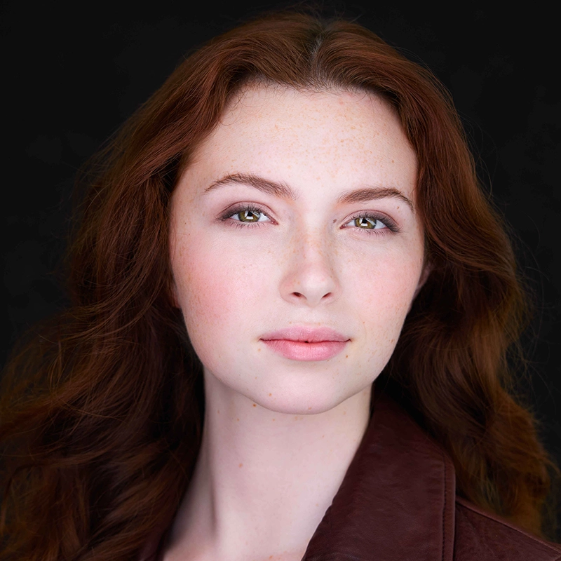 Danielle Serrano-Bremer, a young woman with fair skin, long wavy auburn hair, and light hazel eyes, gazes slightly past the camera with a calm expression. She wears natural makeup and a brownish jacket against a solid black background.