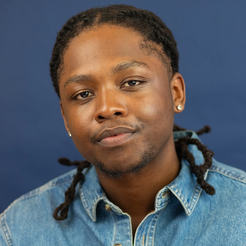 Christian Brailsford, a young man with medium-dark skin, poses in front of a solid dark blue background. He wears a blue denim button-up shirt, has short dreadlocks, a slight smile, and small stud earrings in both ears.