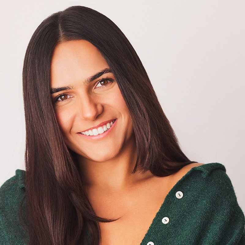 Caroline Bowers, a young woman with long, straight dark brown hair, smiles warmly at the camera. She wears a dark green top with white buttons, one side slightly off her shoulder. The plain light background keeps her as the clear focus.