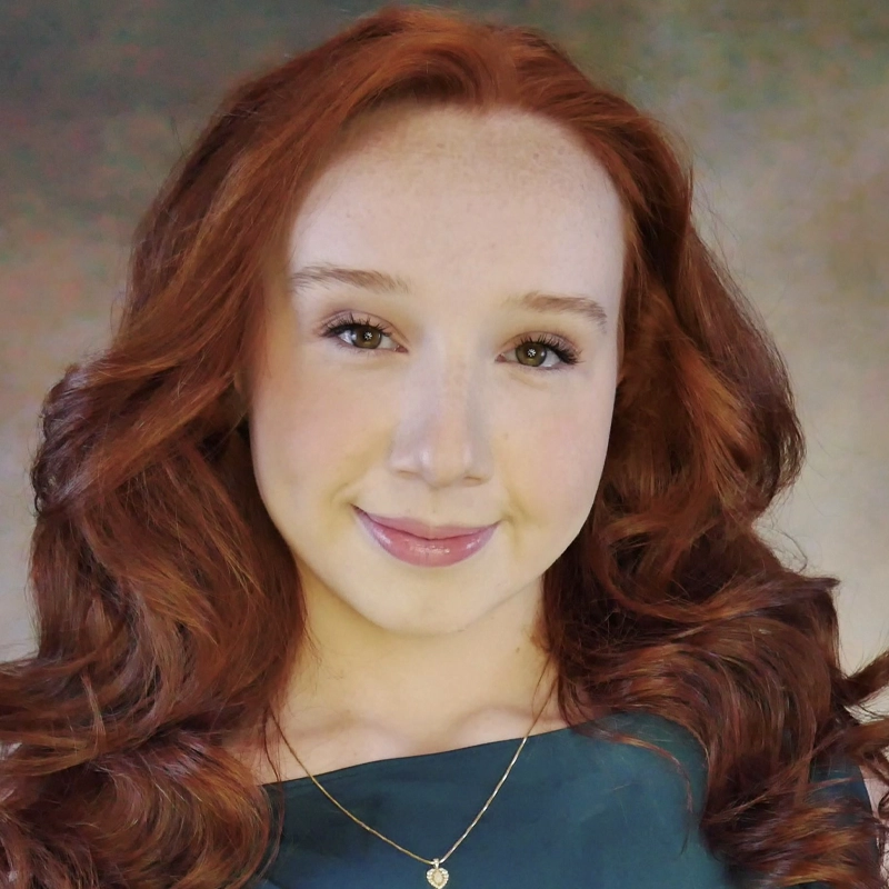 Madison Palmer, a young woman with long, wavy red hair and fair skin, smiles gently. She wears a dark green top and a gold necklace with a small heart pendant. The softly blurred background keeps the focus on her face.