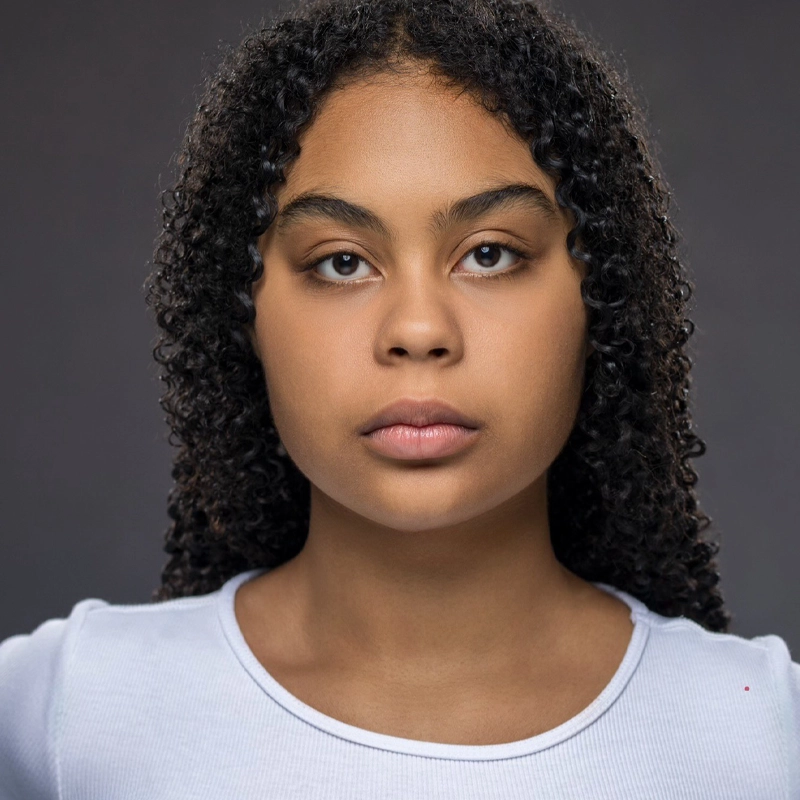 A young person with medium-brown skin and curly dark hair stares directly at the camera with a neutral expression. They are wearing a light blue, scoop-neck shirt. The background is a plain, dark gray, and the lighting is soft and even.