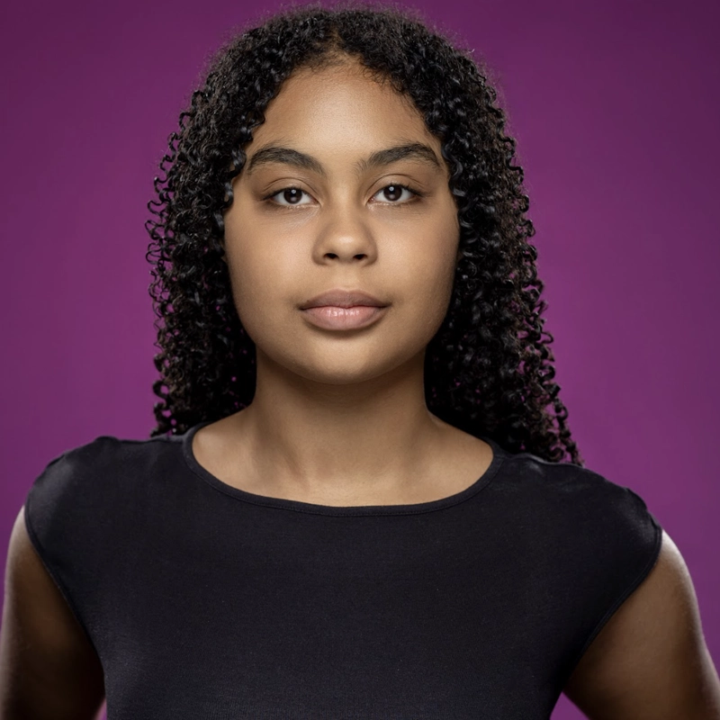 A young woman with curly dark hair, wearing a plain black top, stands confidently against a solid purple background. She looks directly at the camera with a neutral expression, her arms slightly bent at her sides.