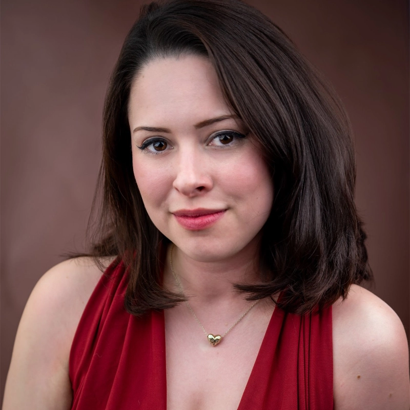 A woman with fair skin and shoulder-length dark brown hair wears a sleeveless red dress and a gold heart necklace. She has natural makeup with pink lipstick and is smiling softly, posing in front of a dark brown, blurred background.