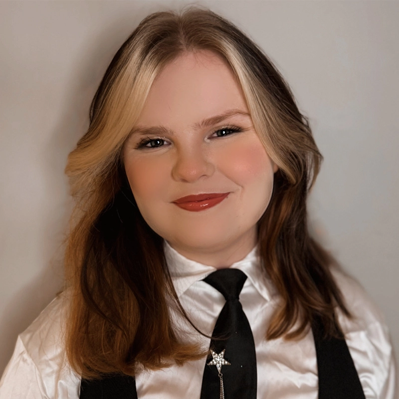Peyton Amaral, with shoulder-length brown hair and blonde highlights, smiles softly. They wear a white shirt, black tie with a star pin, and black vest. The light background adds to the portrait’s professional yet friendly feel.