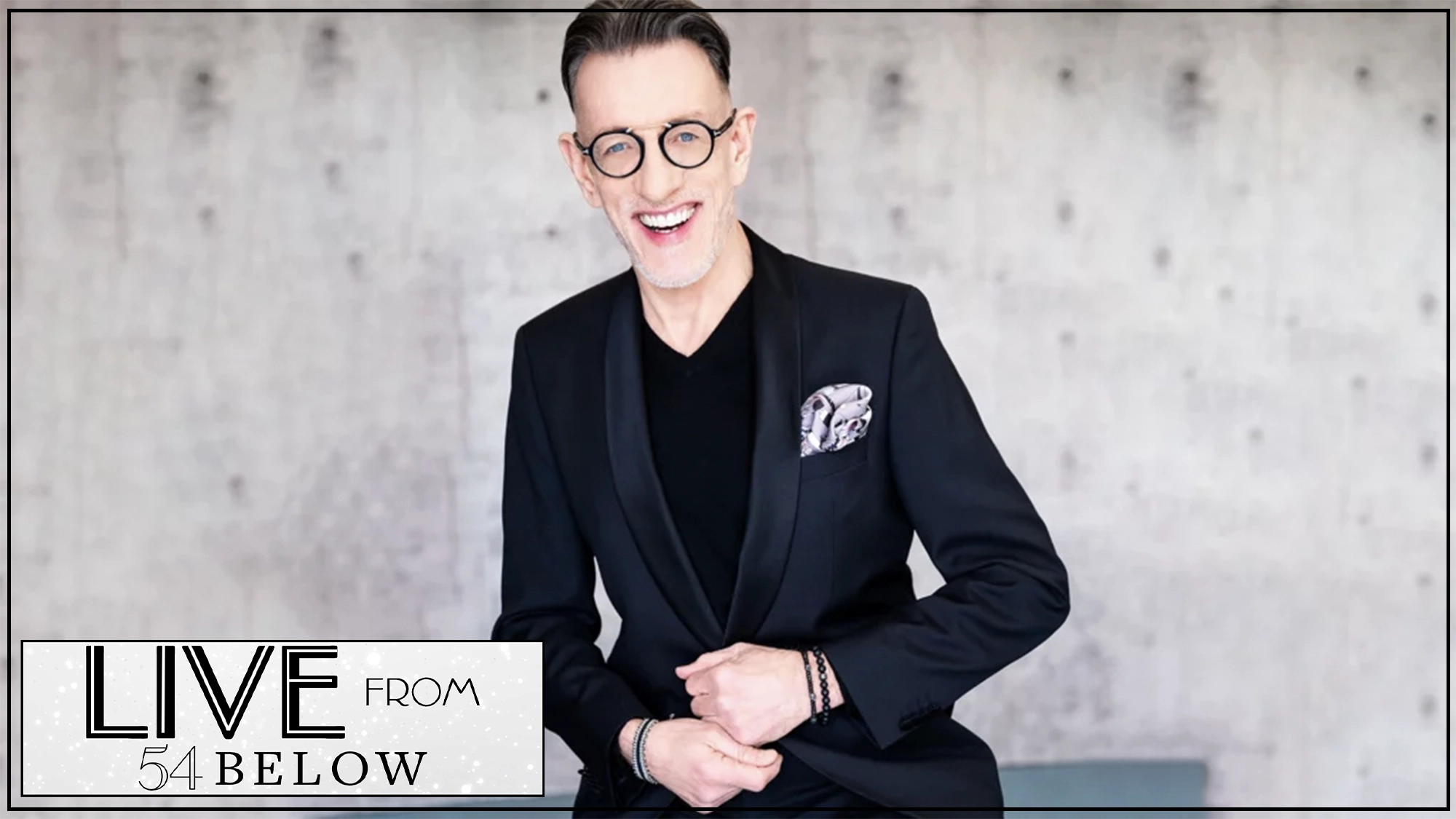 Phillip Officer show page A smiling man with short dark hair, wearing round glasses, a black blazer, and a patterned pocket square, sits posed against a light gray textured wall. The “Live from 54 Below” graphic hints at a special livestream celebrating Mabel Mercer and Phillip Officer.