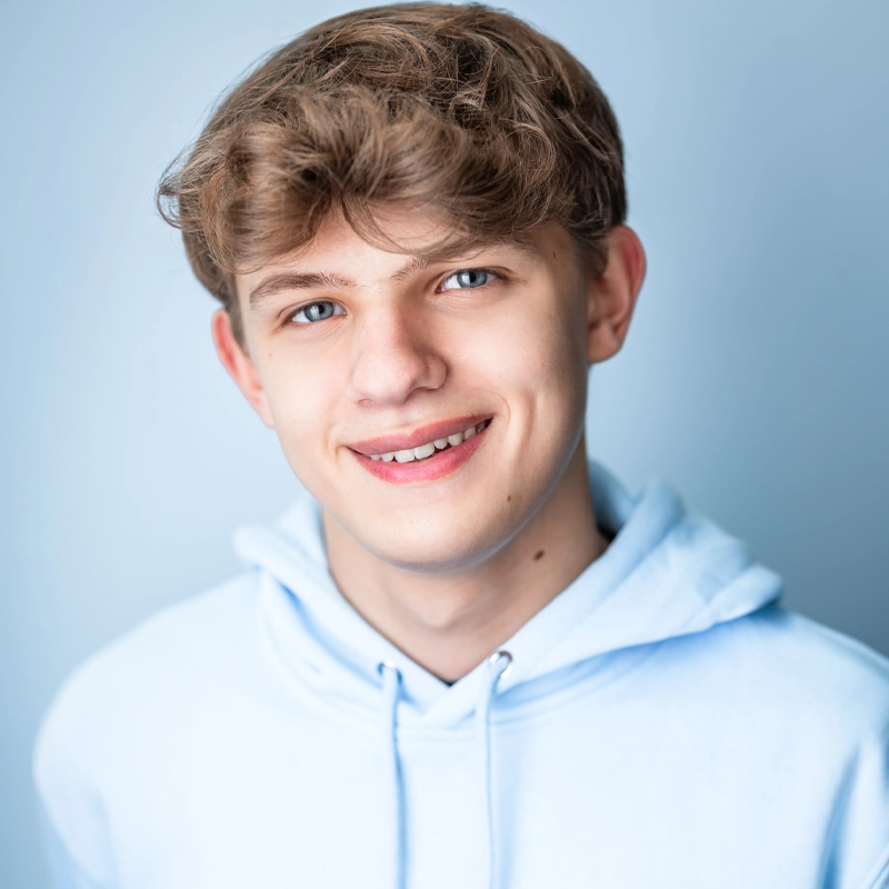 A teenage boy with light brown wavy hair and blue eyes smiles at the camera. Philip Podhurst is wearing a light blue hoodie against a soft blue background. The lighting highlights his clear skin and friendly expression.