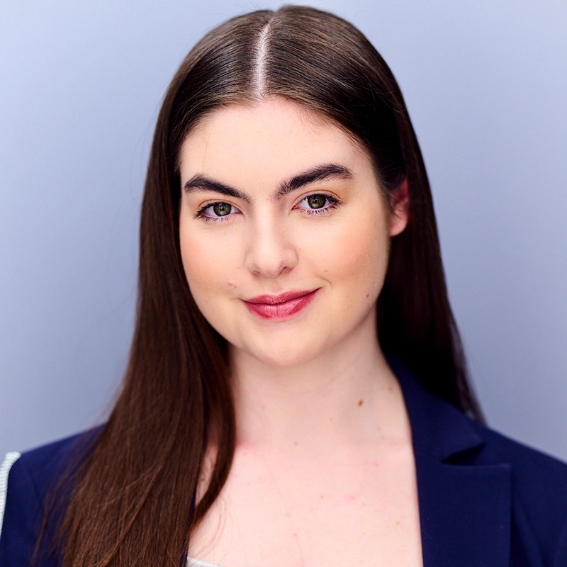 Mickey Wirtz, a young woman with straight, long brown hair and fair skin, smiles softly. She wears a navy blazer over a light top and poses confidently against a plain, light grey background, her expression warm and approachable.