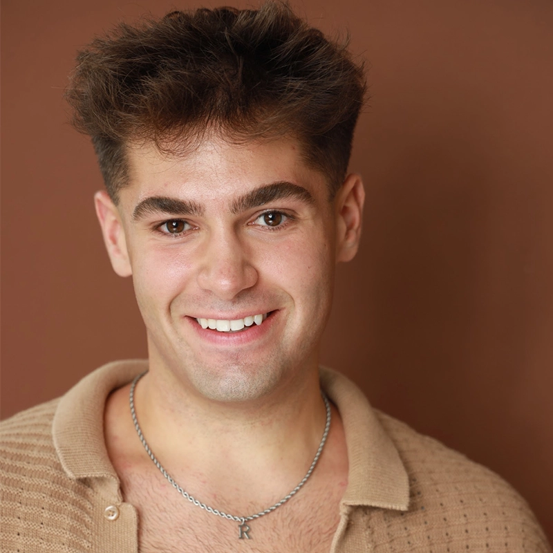 A young man with short, wavy brown hair smiles at the camera. RJ Vandenbrouck wears a beige textured shirt with the collar open and a silver chain necklace with an 