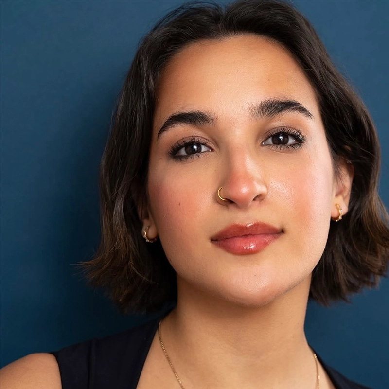 Young person with short, dark hair and a slight smile poses against a solid blue background. They wear a sleeveless dark top, gold hoop earrings, a nose ring, and a gold necklace. Their makeup is natural, with glossy lips and groomed eyebrows—captured in the distinctive style of Michael Mariniello.
