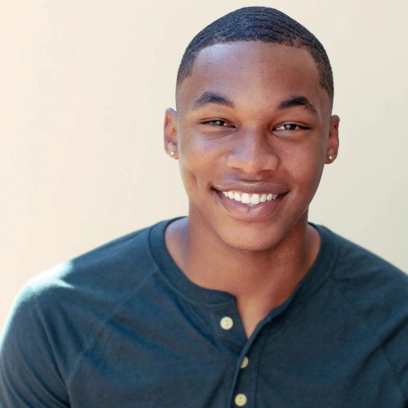 Michael Reed, a young man with short hair and stud earrings, smiles warmly at the camera. He wears a dark, henley-style shirt and is posed against a plain, light-colored background with bright, even lighting for a cheerful portrait.