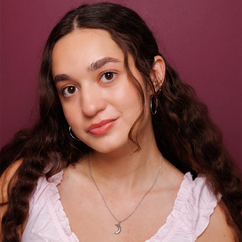 A young woman with long, wavy brown hair wears a pale pink ruffled top, silver hoop earrings, and a silver crescent-moon necklace. She smiles softly against a dark magenta background, looking directly at the camera.