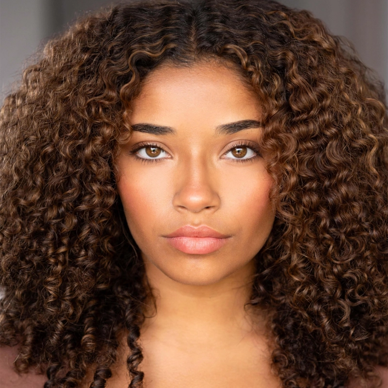 A young woman with medium brown skin and voluminous, dark brown, tight curly hair stares directly at the camera. She has brown eyes, full eyebrows, and natural makeup with soft pink lips, set against a softly blurred background.