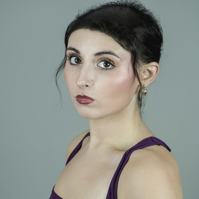 A close-up portrait of Reilly Kearney, a woman with dark hair pulled back, wearing a purple tank top and gold hoop earrings. She is looking directly at the camera with a neutral expression against a plain gray background.