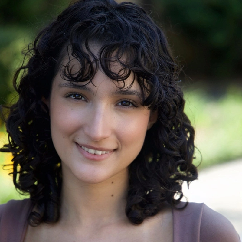 A young woman with fair skin and dark, curly shoulder-length hair smiles gently. She is outdoors, with greenery blurred in the background. She wears a light brown top and natural makeup, giving her a warm and approachable appearance.