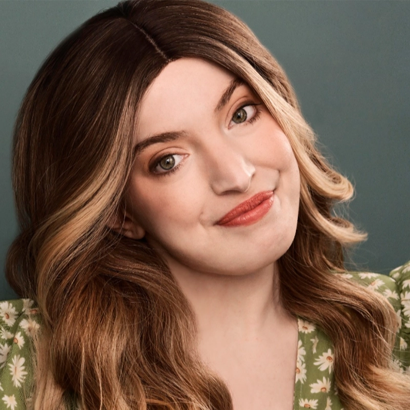 Caroline Weston, a young woman with long, wavy brown hair and fair skin, smiles softly. She wears a green floral dress and natural makeup with rosy lipstick, set against a plain, muted teal background.