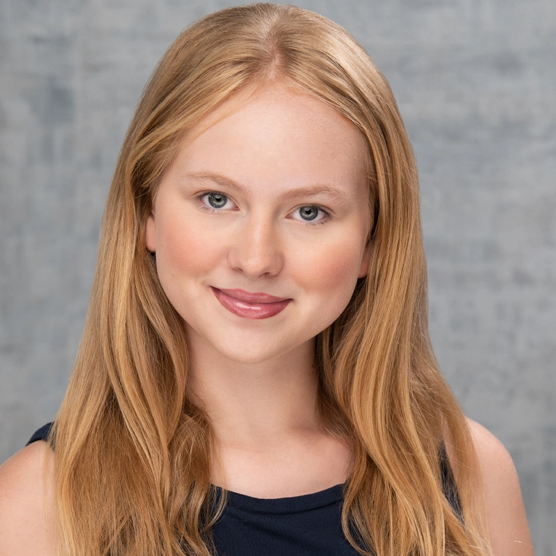 Lilianna Rindal, a young woman with long, straight blonde hair and fair skin, smiles gently at the camera. She has light blue eyes, wears natural makeup and a sleeveless dark top, set against a soft, textured gray background.