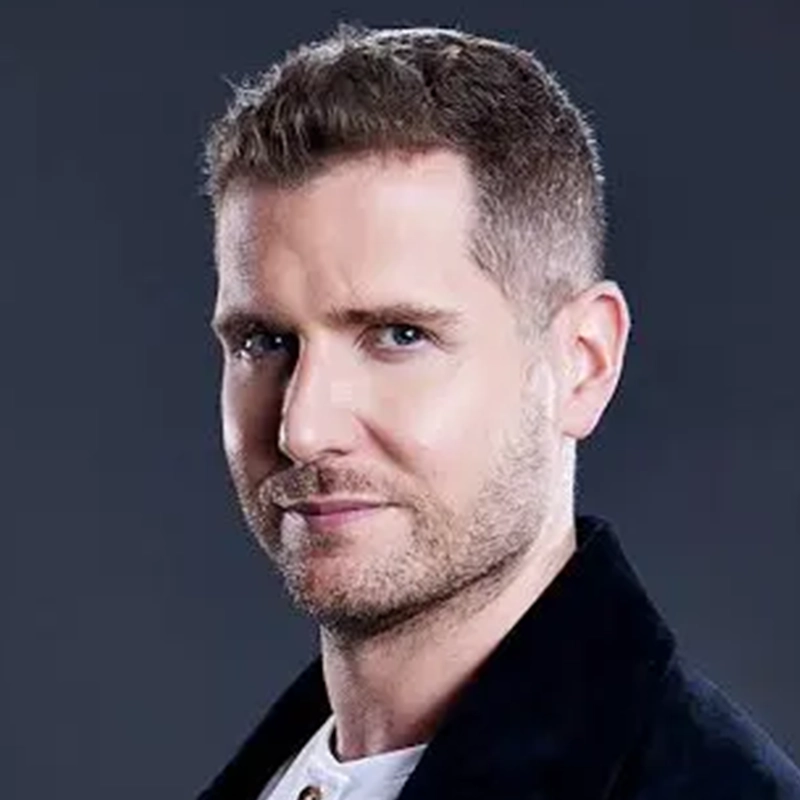 A man with short brown hair, neatly trimmed beard, and light skin looks at the camera with a slight smile. He wears a dark jacket over a white shirt and stands against a plain, dark background.