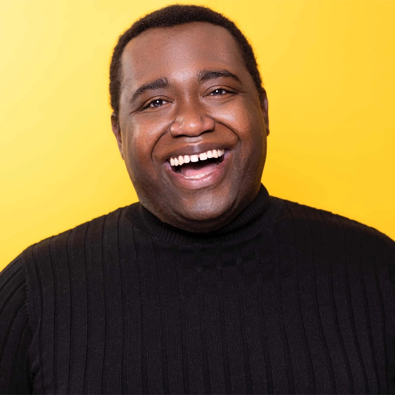 A smiling man, Tyler Price Robinson, wearing a ribbed black sweater stands in front of a solid bright yellow background. He has short black hair and looks directly at the camera with an open-mouthed, joyful expression.