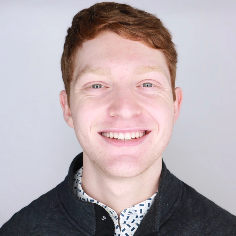 A young man with fair skin and short red hair smiles at the camera. He is wearing a patterned collared shirt under a dark sweater. The background is plain and light gray, giving the image a professional, studio-like feel.