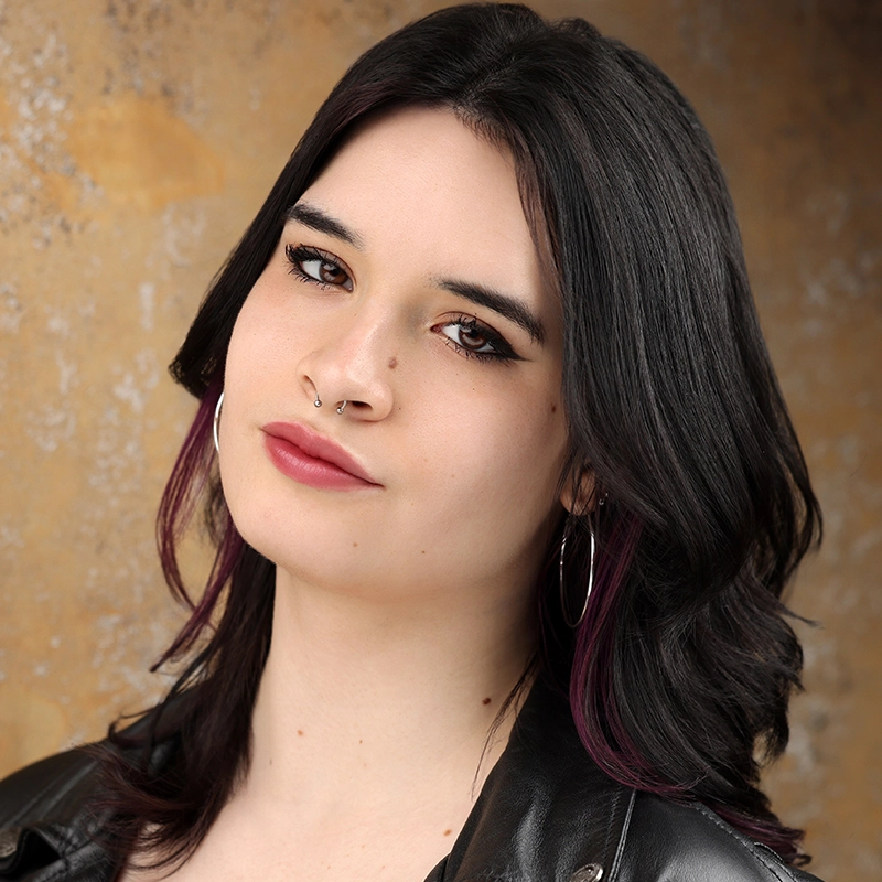 A young woman, Rynn Stephenson, with shoulder-length dark hair with subtle purple tips, wearing hoop earrings, a nose ring, and a black leather jacket, poses in front of a textured light brown background with a soft, confident expression.