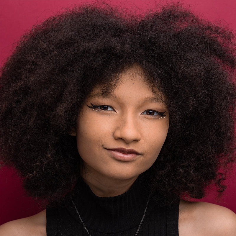 Sahara Williams, a young woman with voluminous curly hair, poses in front of a dark pink background. She wears a sleeveless black turtleneck top, subtle eyeliner, and a slight smile, looking confidently at the camera.