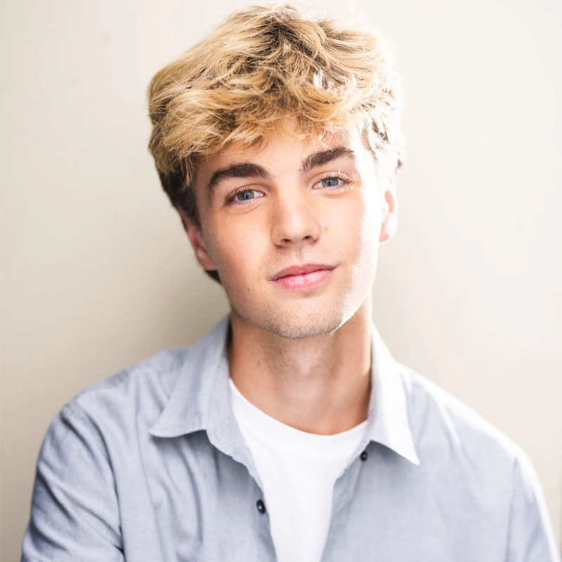 Miles Salerno, a young man with tousled blond hair and blue eyes, smiles softly at the camera. He wears a light blue button-up shirt over a white t-shirt, set against a neutral, softly lit background that creates a warm, inviting feel.