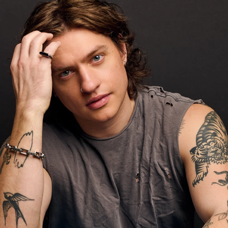A young man with shoulder-length wavy hair and multiple tattoos poses against a dark background. He wears a grey sleeveless shirt and has piercings in his ear and nose.