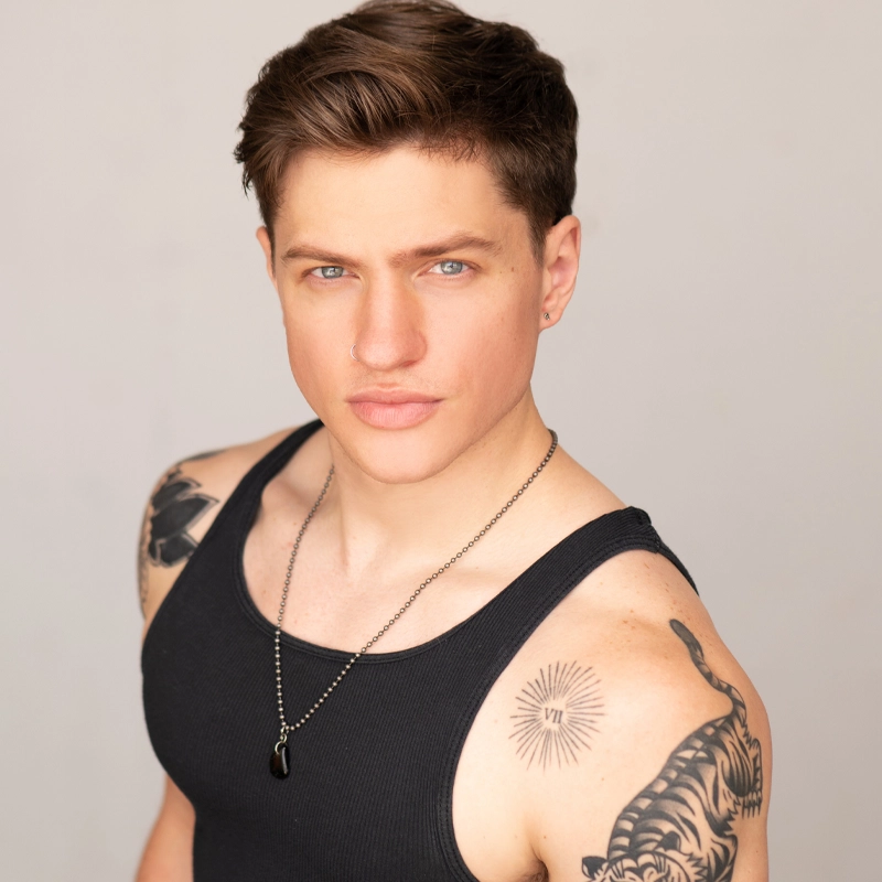 A young man with short brown hair and blue eyes wears a black tank top and a necklace with a dark pendant. He has tattoos on both shoulders—one of a tiger and another resembling rays or a sunburst with Roman numerals. He gazes confidently at the camera.