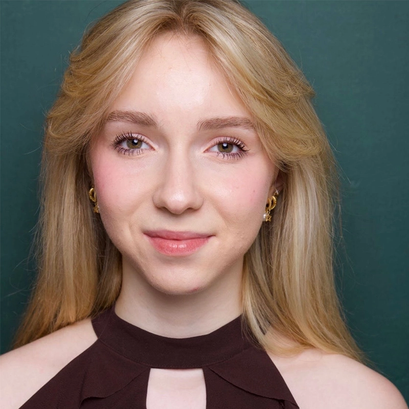 A young woman with long blonde hair smiles softly in front of a dark green background. She wears a sleeveless dark brown top with a high neckline and small gold hoop earrings, styled by RJ Vandenbrouck. Her makeup is natural with light pink lipstick.