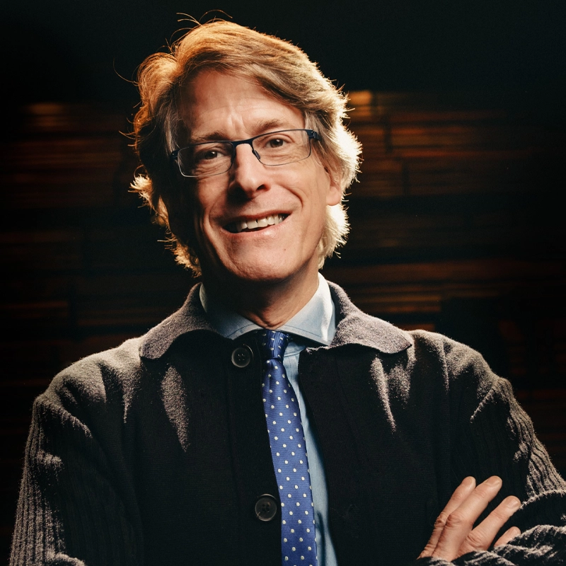 Dick Scanlan, a middle-aged man with light brown hair and glasses, smiles at the camera. He wears a dark cardigan over a collared shirt and blue polka-dot tie, set against dim lighting and wooden panels in the background.