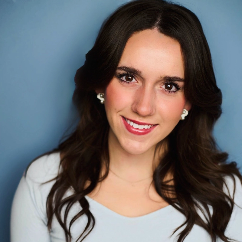 A young woman, Lily Schantz, with long, wavy dark brown hair smiles at the camera. She has brown eyes, light skin, and wears light makeup, pearl earrings, and a light-colored top. The background is a solid blue color.