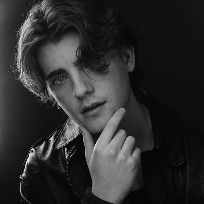 Black and white portrait in the style of Paul Schoeller: a young person with wavy, medium-length hair in a leather jacket, touching their chin thoughtfully while gazing at the camera, softly lit from the left against a dark background.