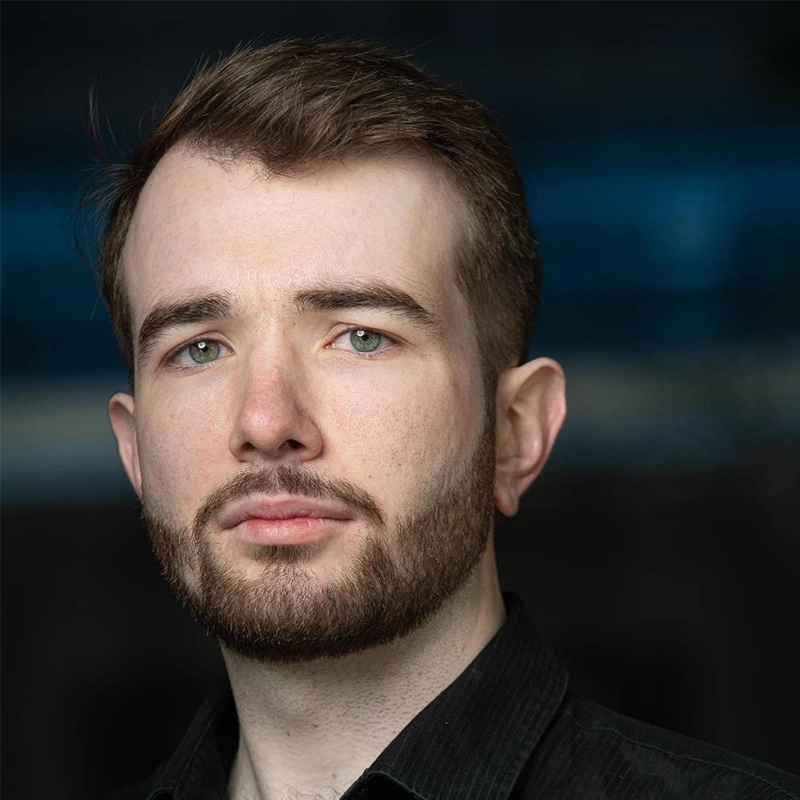 A young man with short brown hair and a neat beard gazes seriously at the camera. He has light skin, blue-green eyes, and is wearing a black collared shirt—a portrait in the distinctive style of Hannah Thompson. The background is dark and blurred.
