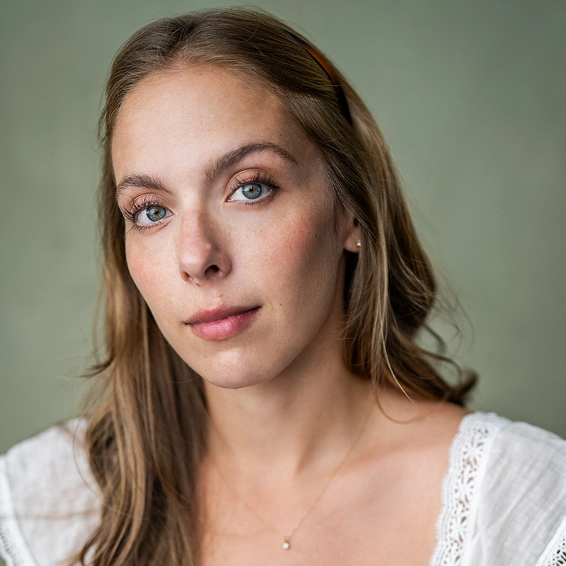 A young woman with long, light brown hair and blue eyes gazes softly at the camera. She wears a white, lace-trimmed top and a delicate gold necklace. The plain, muted green background adds to the calm, natural feel of this Mickey Wirtz portrait.