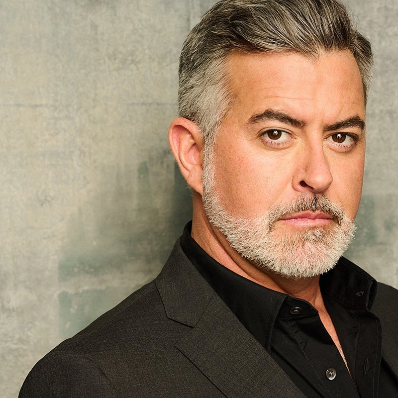 A middle-aged man with gray hair and a trimmed beard wears a dark suit jacket and black shirt. He has a serious expression and is posed against a textured gray background, looking directly at the camera.