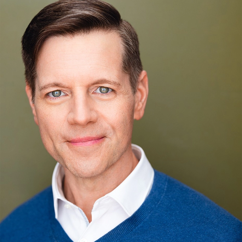 Billy Sharpe, with neatly combed brown hair, blue eyes, and light skin, smiles gently. He wears a white collared shirt under a blue sweater. The muted olive green background and bright lighting highlight his face.