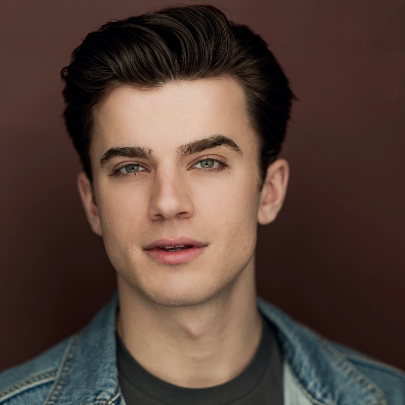 A young man with fair skin, short dark brown hair, and green eyes looks at the camera. He wears a denim jacket over a dark shirt, and the background is a solid, muted reddish-brown color. Scott Silagy is softly smiling with a relaxed expression.