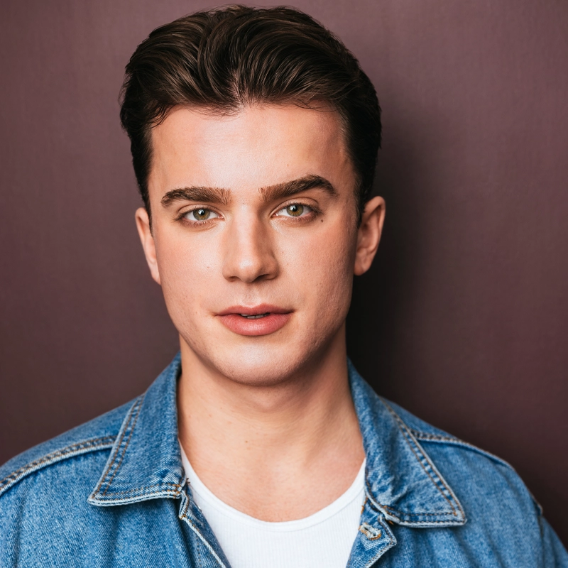 Scott Silagy, a young man with short brown hair and green eyes, stands against a plain dark purple background. He wears a blue denim jacket over a white t-shirt and looks directly at the camera with a neutral expression.