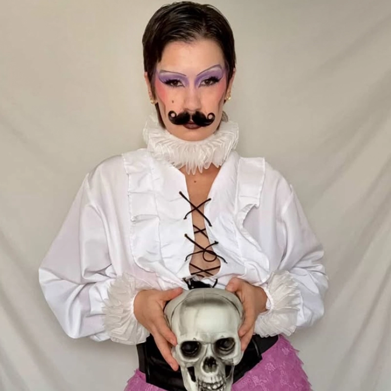 A person with dramatic purple eye makeup, drawn-on curled mustache, and slicked-back hair wears a white ruffled shirt with black laces and a lavender skirt. They hold a realistic human skull and stand against a plain light background.