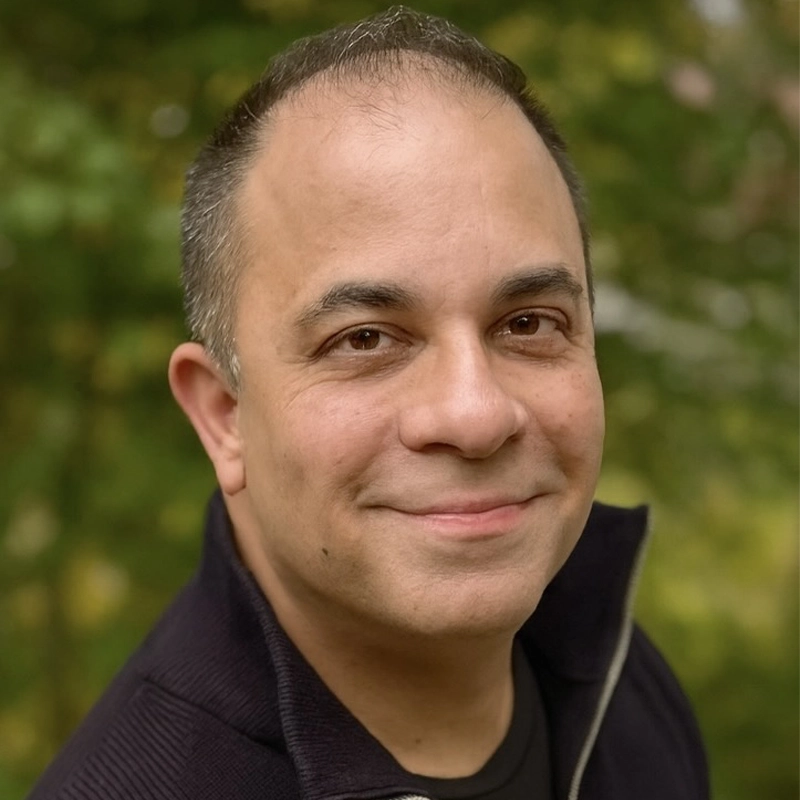 A middle-aged man with short dark hair and a receding hairline smiles softly at the camera. He wears a dark zip-up sweater over a black shirt. The background is blurred greenery, suggesting an outdoor setting.