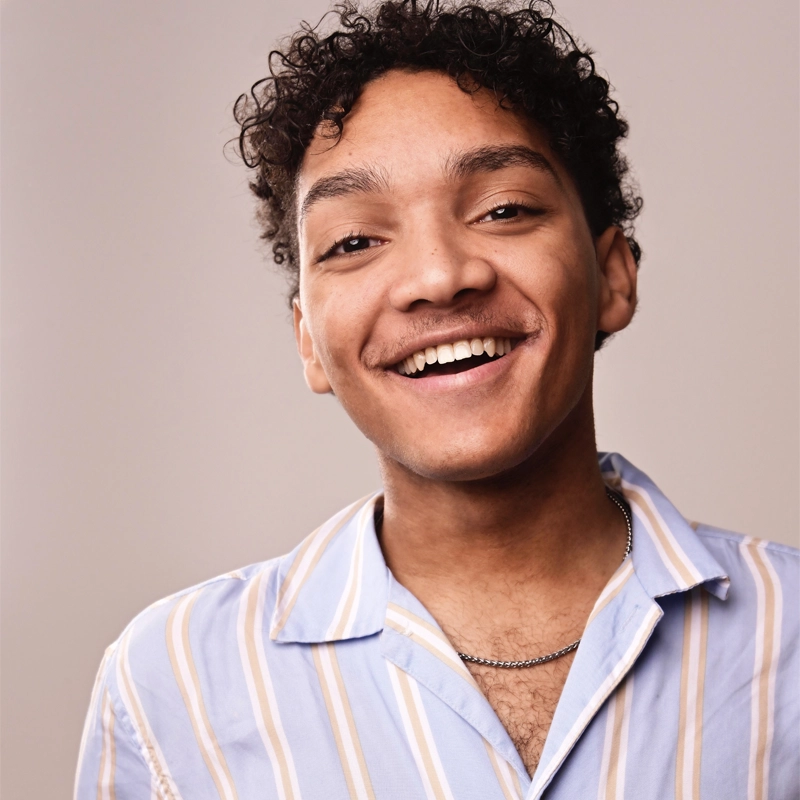 A young man with short curly hair, Gavin Lee Smith, smiles warmly at the camera. He wears a light blue shirt with beige and white stripes and a thin chain necklace. The softly lit background creates a friendly and inviting atmosphere.