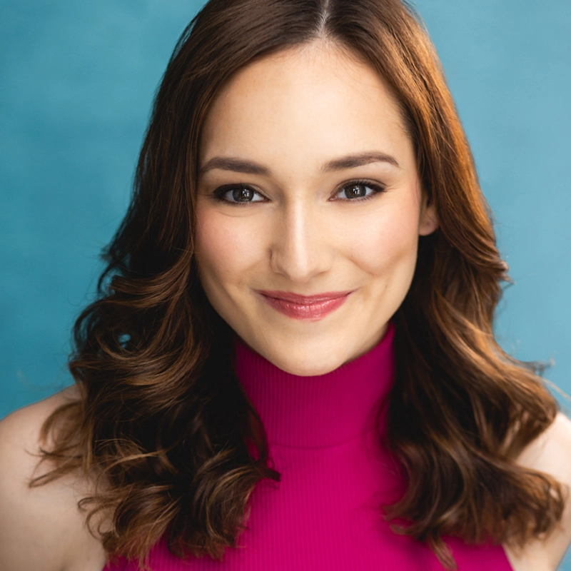 Taryn Smithson, a young woman with wavy brown hair, wears a sleeveless magenta turtleneck top and smiles gently at the camera. She has light skin, natural makeup, and stands against a solid teal blue background with bright, even lighting.