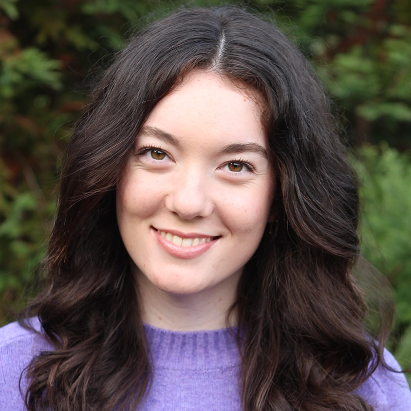 A young woman with long wavy brown hair smiles gently at the camera. Mickey Wirtz is wearing a purple sweater and is outdoors, with blurred green foliage in the background. The natural lighting highlights her features.