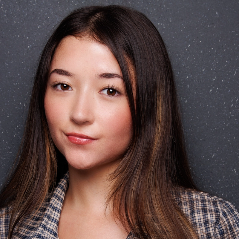 Kenzie Woolridge, a young woman with long, straight brown hair and fair skin, poses in front of a dark, textured background. She wears a plaid shirt and has a calm, neutral expression with a slight smile and naturally fresh makeup.