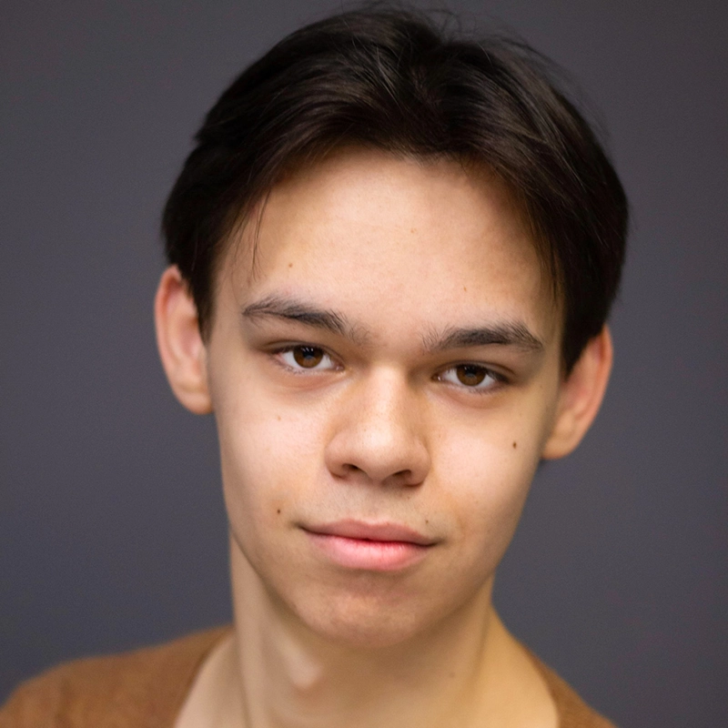 A young person with straight, dark brown hair parted down the middle, light skin, and brown eyes looks directly at the camera with a slight, neutral smile. The background is plain gray, and Danielle Serrano-Bremer wears a light brown shirt.